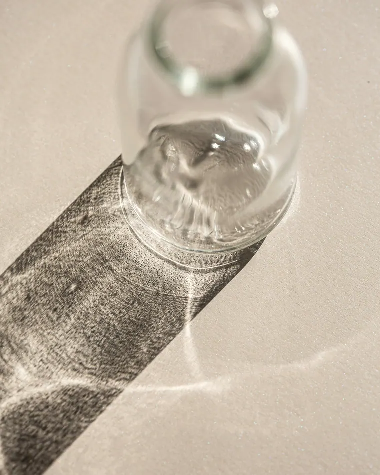 Pote de vidro sobre uma superfície bege, com a sombra atravessada por reflexos de luz.